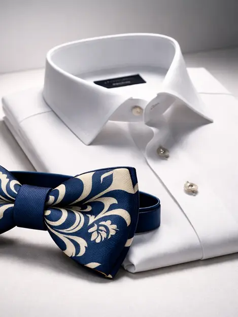 Urban Arabesque navy and ivory pre-tied butterfly bow tie in silk placed beside a folded white dress shirt under sculpted atelier lighting