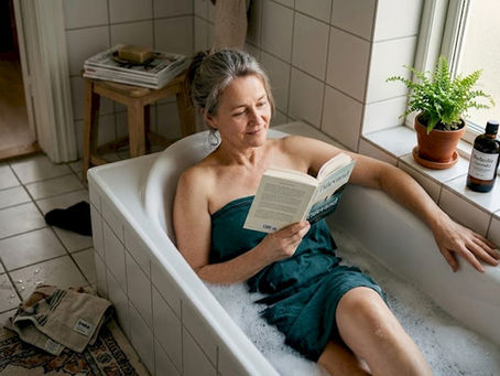 En kvinde slapper af i badekarret med et godt blad, mens solens bløde stråler falder ind gennem vinduet.