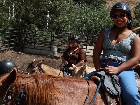 Person horseback riding outdoors, wearing helmet and sunglasses, with others riding nearby.