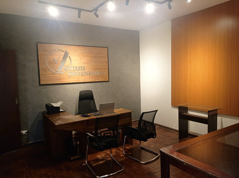 Foto do escritório da Verum Engenharia, onde se observa uma mesa com duas cadeiras, um notebook a logo da empresa. O chão e a porta são de madeira.