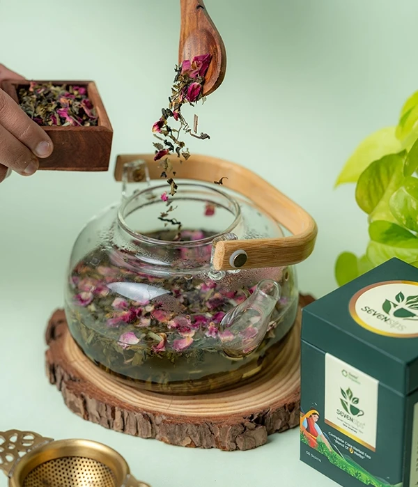 Hands pour dried herbal tea leaves into a glass teapot on a wood slice beside a Seven Sips tea box, with a mint green backdrop.