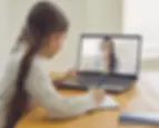 Menina com trança assistindo a uma aula online no notebook, interagindo com a professora em chamada de vídeo.