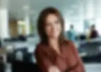 Mulher de cabelos castanhos, vestindo blusa marrom, sorrindo com os braços cruzados em um ambiente corporativo.
