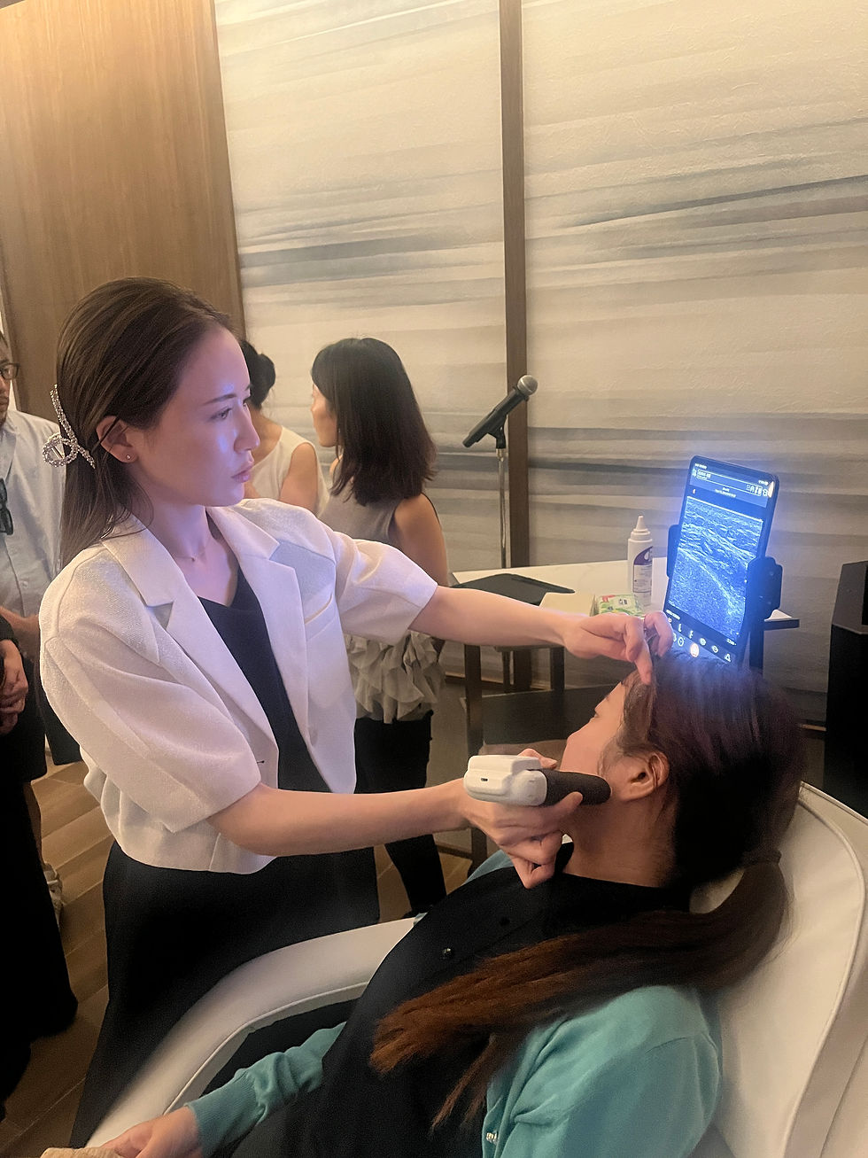 Woman performs a facial ultrasound scan on a seated patient in a clinic, with a monitor and microphone nearby.
