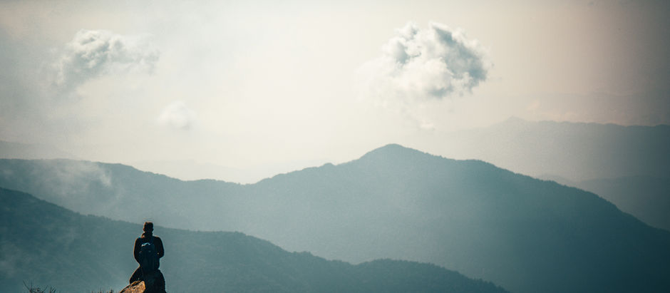 Lone hiker with backpack sits on a rocky cliff, overlooking misty blue mountain layers and clouds at sunrise.