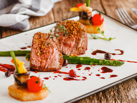 Grilled steak with herbs on asparagus, cherry tomatoes, and decorative sauce on a white plate, set on a rustic wooden table.
