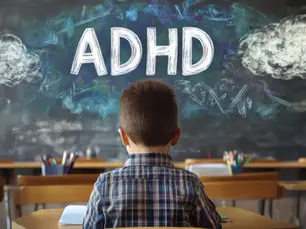 Child in plaid shirt sits at a desk in a classroom. "ADHD" is written on the blackboard with colorful chalk clouds. Classroom setting.