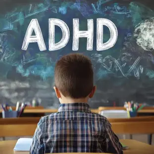 Child in plaid shirt sits at a desk in a classroom. "ADHD" is written on the blackboard with colorful chalk clouds. Classroom setting.