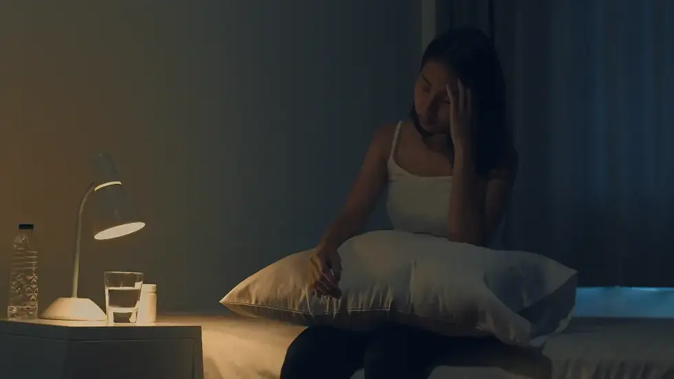 A woman sits on her bed with a troubled expression, holding a pillow, as soft lamp light illuminates the room, evoking the theme of overthinking at night.