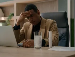 A man sits at his desk, visibly stressed, resting his head in his hand while gazing at a laptop. A glass of water and some papers lie beside him, hinting at a challenging day at the office.