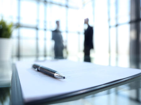 A close-up of a pen resting on a signed contract on a glass table, with two blurred business professionals standing in a modern office background.