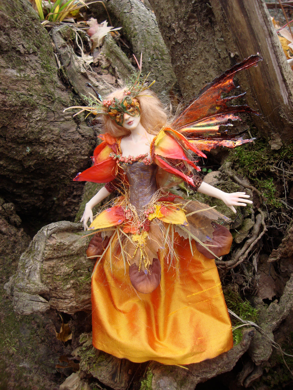 a blond fae with orange fallings, and an orange dress fringed with fall leaves and fiber, a rust colored corset. she is wearing an antlered mask made of fall leaves. outside by a tree
