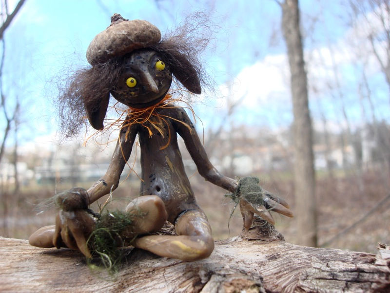 a round faced thin goblin with brown skin and yellow eyes, droopy rabbit ears. seated cross legged. he wears an acorn cap as a hat.
