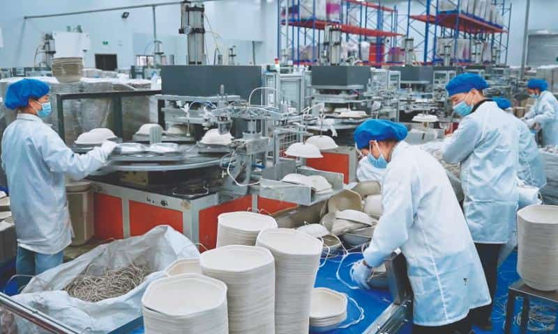 Workers in white coats, blue hairnets and face masks operating machinery and stacking white circular bagasse tableware products on a factory production line.