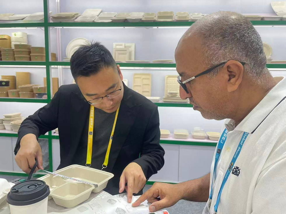 Two businessmen examining and measuring a beige biodegradable food container with a measuring tape at a trade exhibition booth. The background shows shelves filled with eco-friendly disposable tableware.