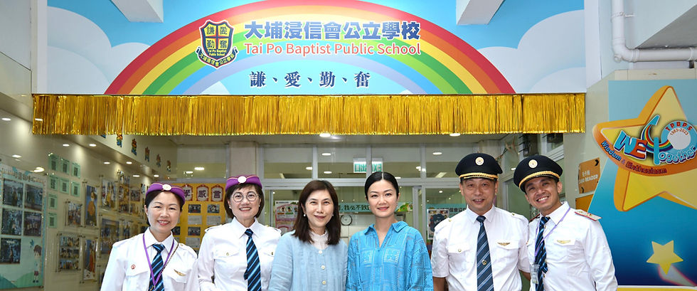 六位嘉賓與教職員在大埔浸信會公立學校校門口合影,門楣上方有彩虹與校徽圖案,寫著「謙・愛・勤・奮」。其中兩位女性穿著紫色帽子與白色制服、兩位男性穿著飛行員制服,另有兩位嘉賓身著便裝站於中間,背景可見校園走廊與牆上展示的照片與作品。