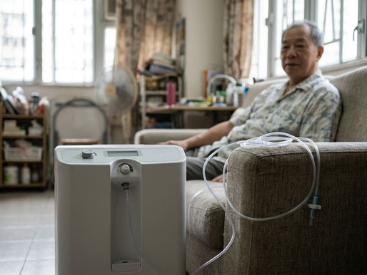 長者伯伯坐在家居客廳沙發上,身旁放著一部白色機身的家用氧氣機,鼻導管垂放在扶手上,畫面對焦在機身及鼻導管,人物臉部因景深稍為虛化
