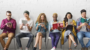 Six young adults sit on chairs against a white brick wall, reading on tablets, phones, and books, smiling and relaxed.