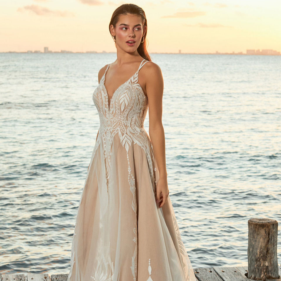 A girl standing in front of an ocean wearing a mocha and ivory wedding dress with boho lace sequin detailing