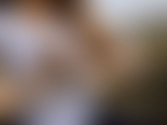 Woman in white tank top doing yoga, hands in shakti mudra. Long hair, rings visible. Blurred outdoor background, calm mood.
