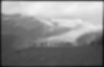 Archival image. Line of hikers with walking poles on a rocky ridge above a glacier and snowy peaks in a black-and-white mountain landscape.