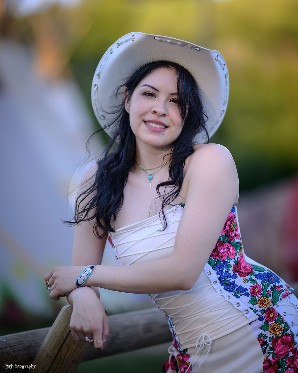 Artist Kristy North Peigan. Smiling woman in a white cowboy hat and floral dress leaning on a wooden fence outdoors; @rjvfotography watermark.