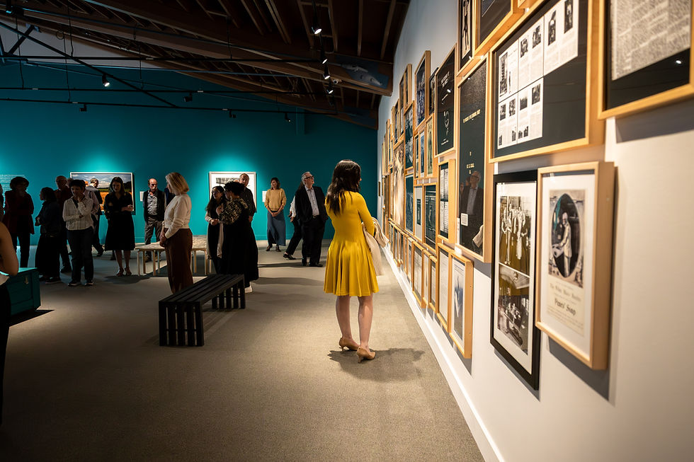 Visitors browse a museum gallery with teal walls at The Whyte for the Thick as Thieves exhibition; a woman in a yellow dress studies a wall of framed exhibits.