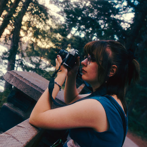 Woman with glasses photographs outdoors near a stone structure in a forest, holding a camera up with a focused expression.