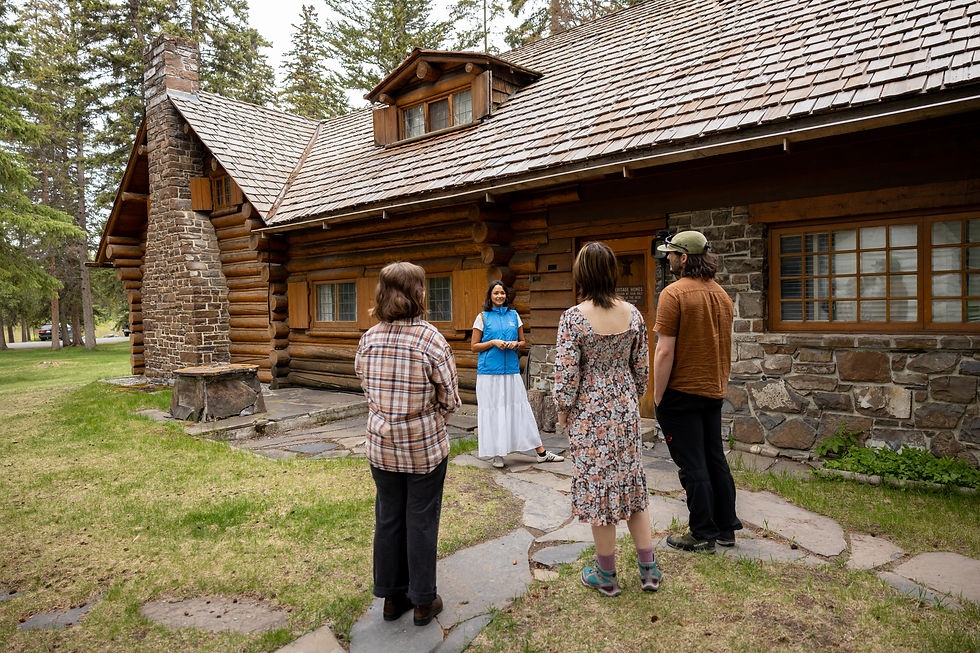 Tour guide speaks to four visitors outside a rustic log cabin with stone chimney, surrounded by trees, on a guided Historic Homes Tour with The Whyte in Banff, Alberta.