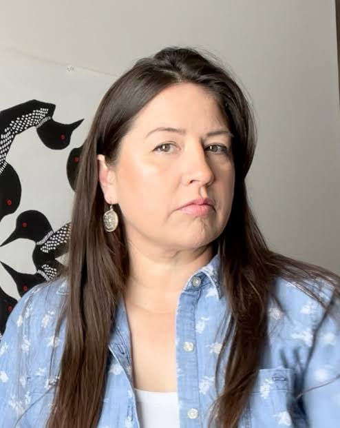 Artist Hali Heavy Shield. Woman in a blue patterned shirt looks off-camera indoors, with black bird wall art behind her and a neutral expression.