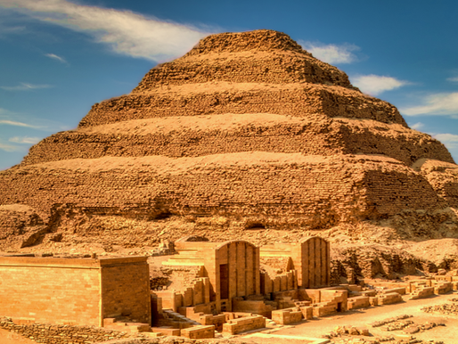 A large stepped stone pyramid rises from the desert landscape under a blue sky with wispy clouds. The ancient structure, known as the Step Pyramid of Djoser, has six distinct layers and is made of weathered limestone blocks. In front of the pyramid are partially restored stone buildings and ruins, part of the surrounding mortuary complex at Saqqara, Egypt.