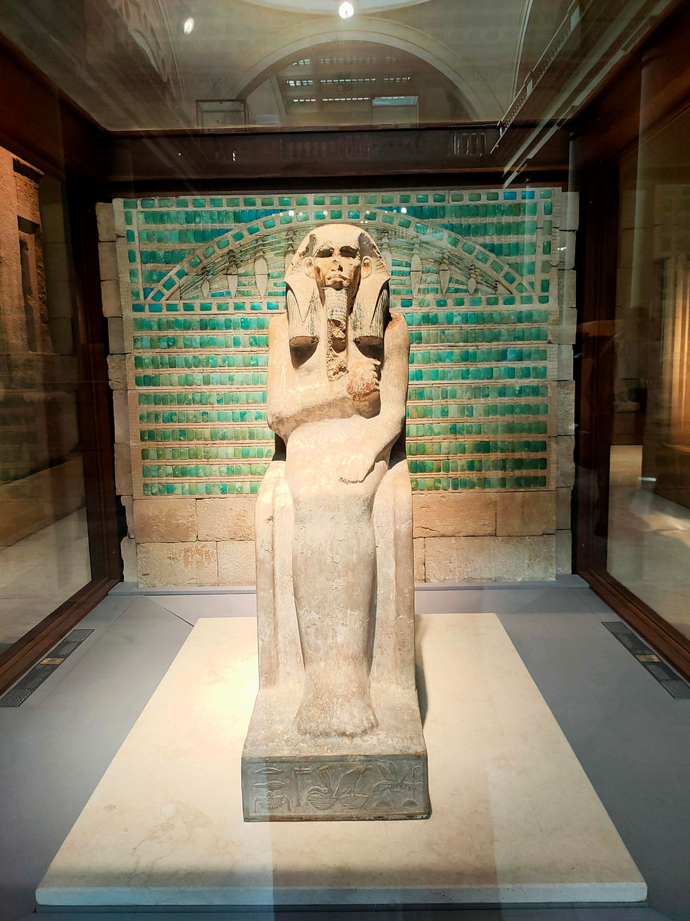A seated limestone statue of Pharaoh Djoser displayed in a glass case at the Egyptian Museum in Cairo. The pharaoh wears a royal headdress and false beard, with arms resting on his thighs. Behind the statue is a reconstructed wall made of turquoise-colored faience tiles, framed by a stone arch.