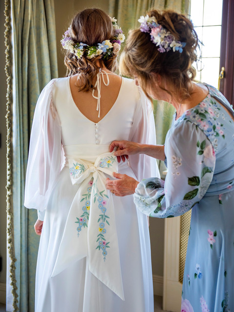 Wildflowers embroidered onto bow of bride's dress. Bride faces away from the camera whilst Mother adjusts the bow. Bow features small embroidered flowers.