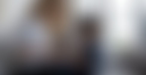 Woman and child on a sofa, woman talking with hands open, child looking down, appearing thoughtful or sad, large windows in the background.