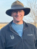 Man smiling, wearing glasses, a wide-brim hat, and a gray Columbia jacket. Outdoor setting with bare tree branches in the background.