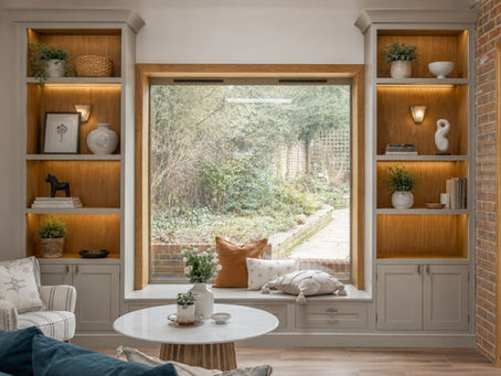 Large oak-framed picture window with bespoke painted joinery, illuminated display shelving, and built-in window seat styled with layered neutral cushions overlooking a soft landscaped garden.