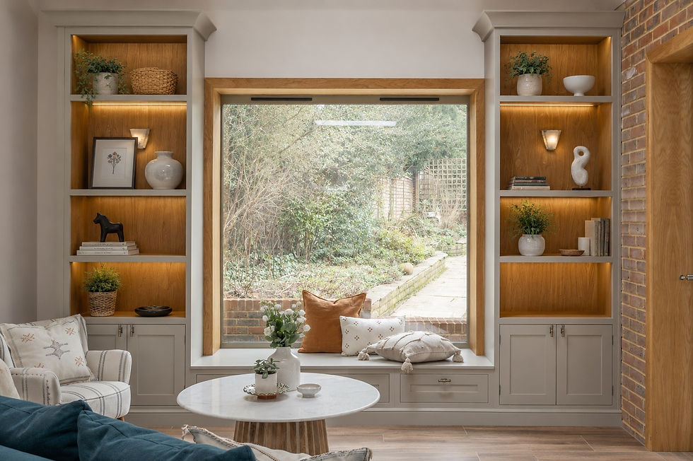 Large oak-framed picture window with bespoke painted joinery, illuminated display shelving, and built-in window seat styled with layered neutral cushions overlooking a soft landscaped garden.