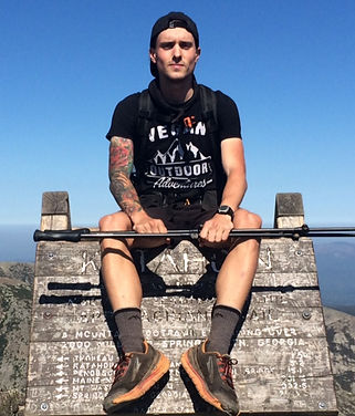 Colton on top of a wooden sign at the top of Katahdin, a mountain in Maine that marks the end of the Appalachian Trail, he is wearing a shirt that says "Vegan Outdoor Adventures"