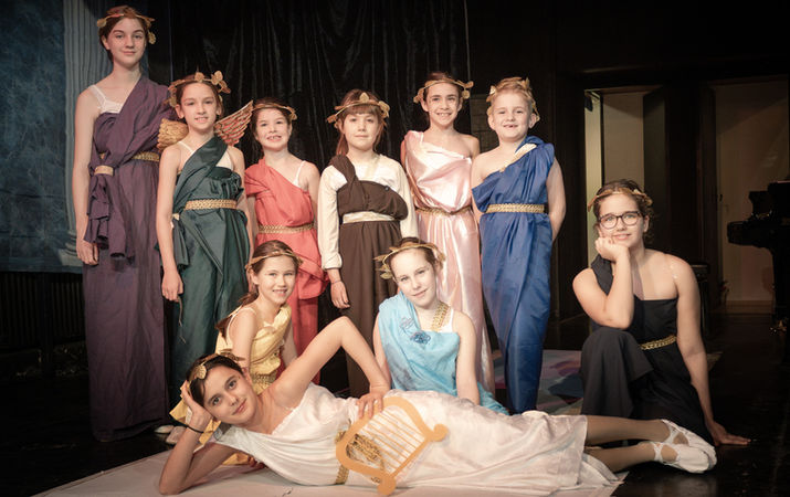 Gyerekek görög jelmezekben pózolnak egy színpadon, egyikük lantot tart.