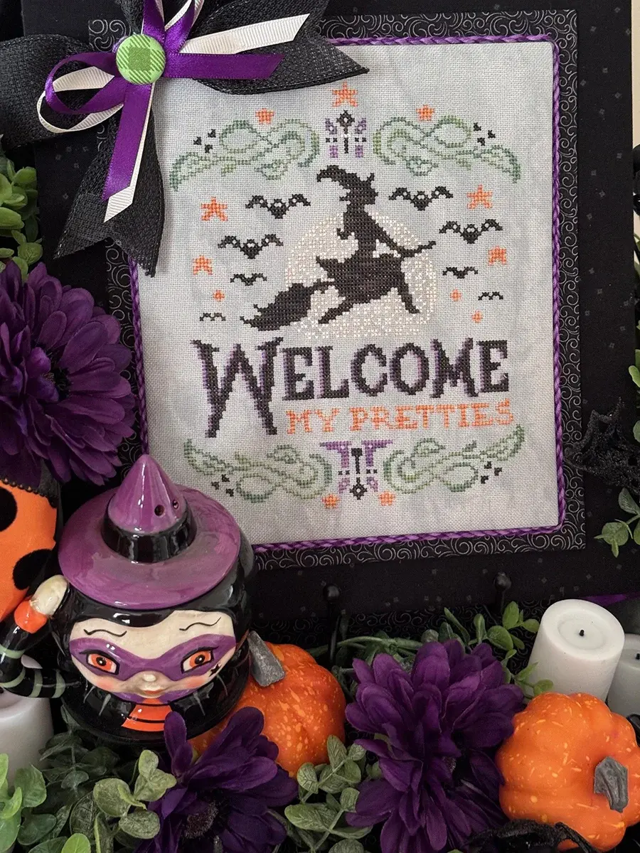 Halloween counted cross stitch pattern with a witch. There is a spooky green border with purple candles and orange stars. The witch is flying in front of a full moon, and she is surrounded by bats. The text is stitched with a spooky font that says "Welcome, My Pretties". The x stitch pattern is stitched with green, orange, black, and purple embroidery thread. The cross stitch finish is displayed on a gray evenweave cloth and has a black fabric frame. It is surrounded in purple flowers, pumpkin picks, eucalyptus, LED candles, and a witch ceramic piece.