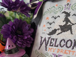Halloween cross stitch display idea. There is a witchy cross stitch pattern, purple flowers, faux greenery, and a ceramic witch.