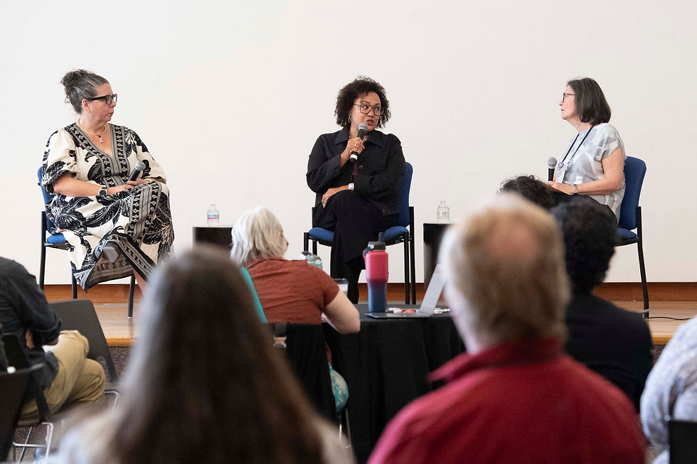 Three women sitting on a stage in front of a white wall, the woman in the middle speaking into a microphone while the other two listen. Attentive audience in the foreground of the photo listens and takes notes