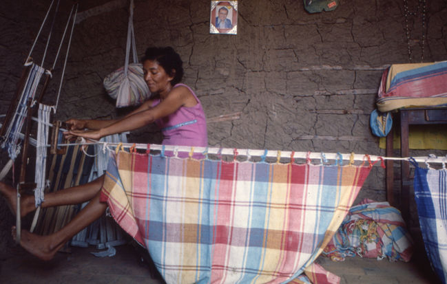Fotografia colorida de uma mulher negra manipulando um tear. Ela tem cabelos escuros curtos, usa uma regata lilás com detalhe em azul. Seus braços e pernas estão esticados para o lado esquerdo da imagem, e cada um está segurando um pedaço de fio de algodão. À frente da imagem uma rede xadrez começa a tomar forma. Aos fundos, parede de barro e madeiras, pedaços de redes espalhadas pelo espaço, um móvel de cor marrom envelhecido, e suportes em madeira utilizados no tear.