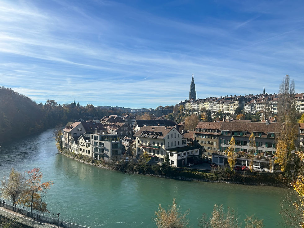 Photo taken by Ames Sawyer. A scenic view of the city of Bern, Switzerland, taken from above the Aare River on a clear autumn day. The turquoise river curves gently through the foreground, lined with rows of houses and small apartment buildings with red and brown rooftops. In the distance, the tall Gothic tower of Bern Minster rises above the historic old town skyline.