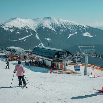 A skier in a pink jacket over looking the top of a chair lift with snowy mountains in the background.