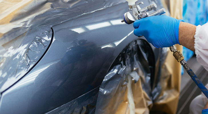 A person wearing blue gloves spray-paints a car's metallic gray surface.