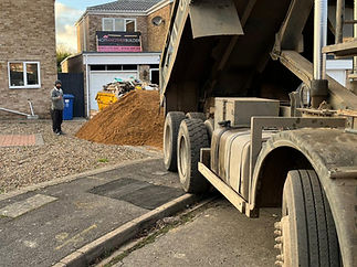 A dump truck unloads soil onto a driveway beside two brick houses