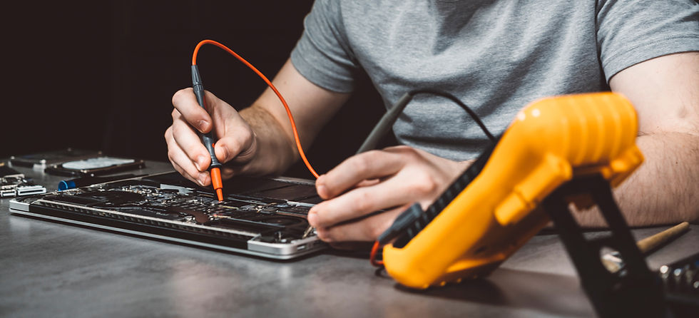 A person in a gray shirt works on a circuit board with a multimeter on a table, focusing intently