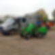 A man sits on a green compact loader with a fork attachment, parked near a silver van and an orange wood chipper