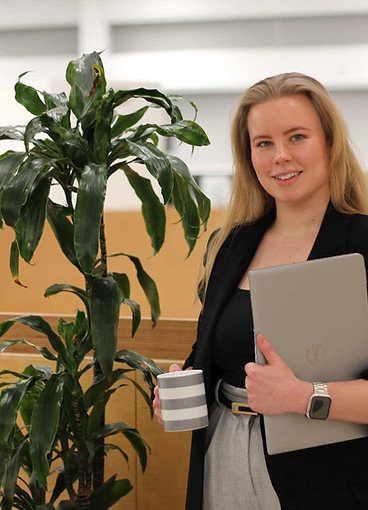 A woman in a black blazer stands next to a plant in an office